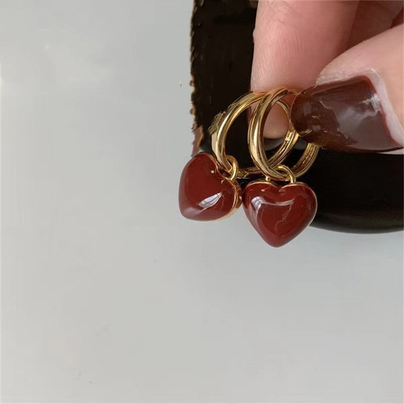 Gold hoop earrings with red heart charms held by a hand against a neutral background