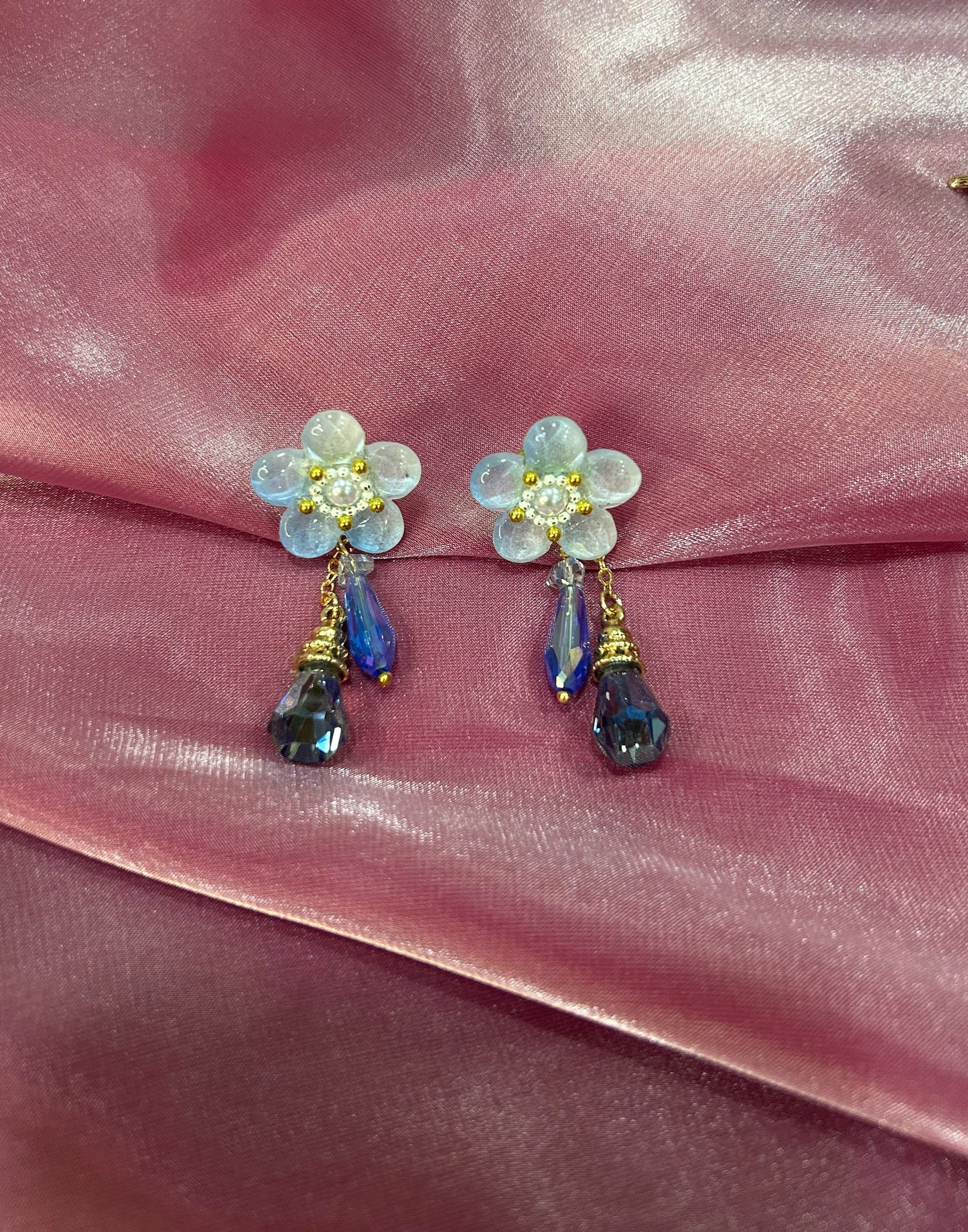 Flower-shaped earrings with blue teardrop charms on a pink fabric background
