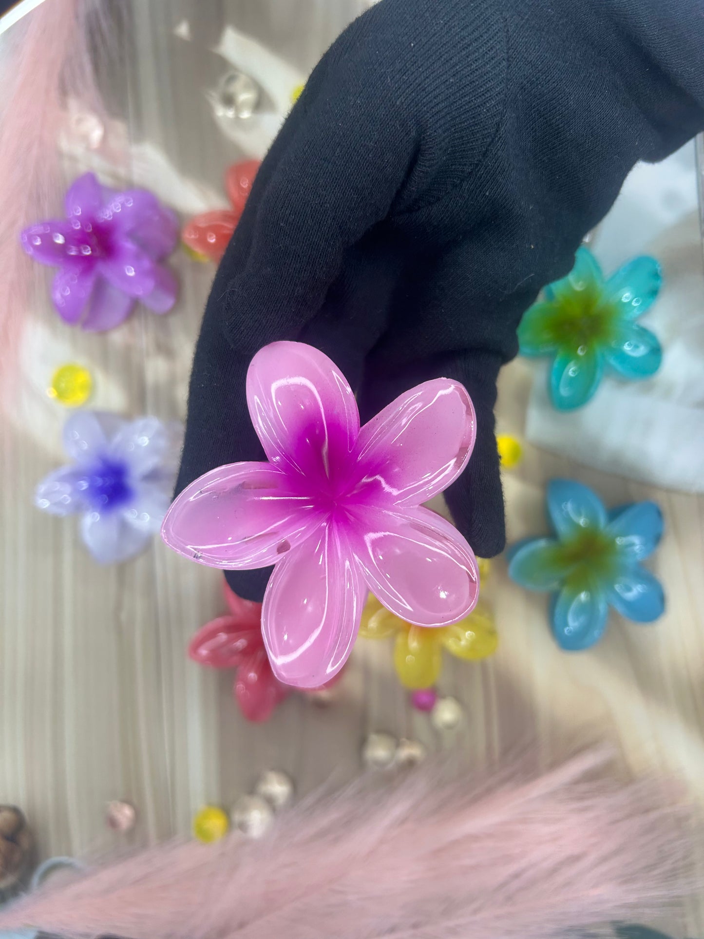 Pink flower-shaped hair clip held by a gloved hand with colorful flowers in the background.