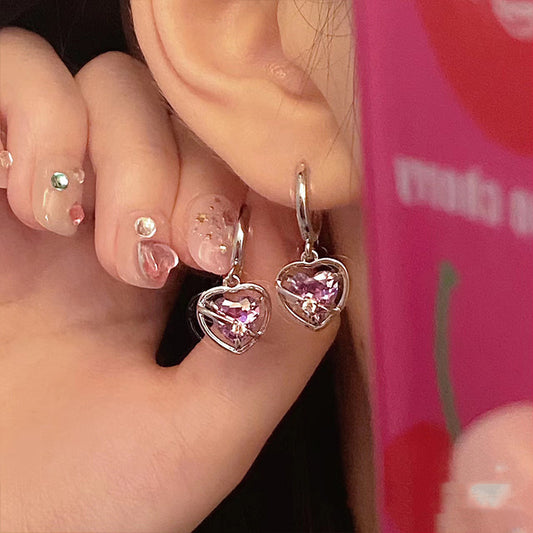 Heart-shaped earrings with pink gemstones worn by a person against a pink background.