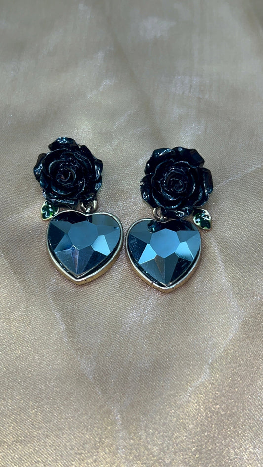 Pair of earrings with black rose designs and blue heart-shaped stones on a beige fabric background.
