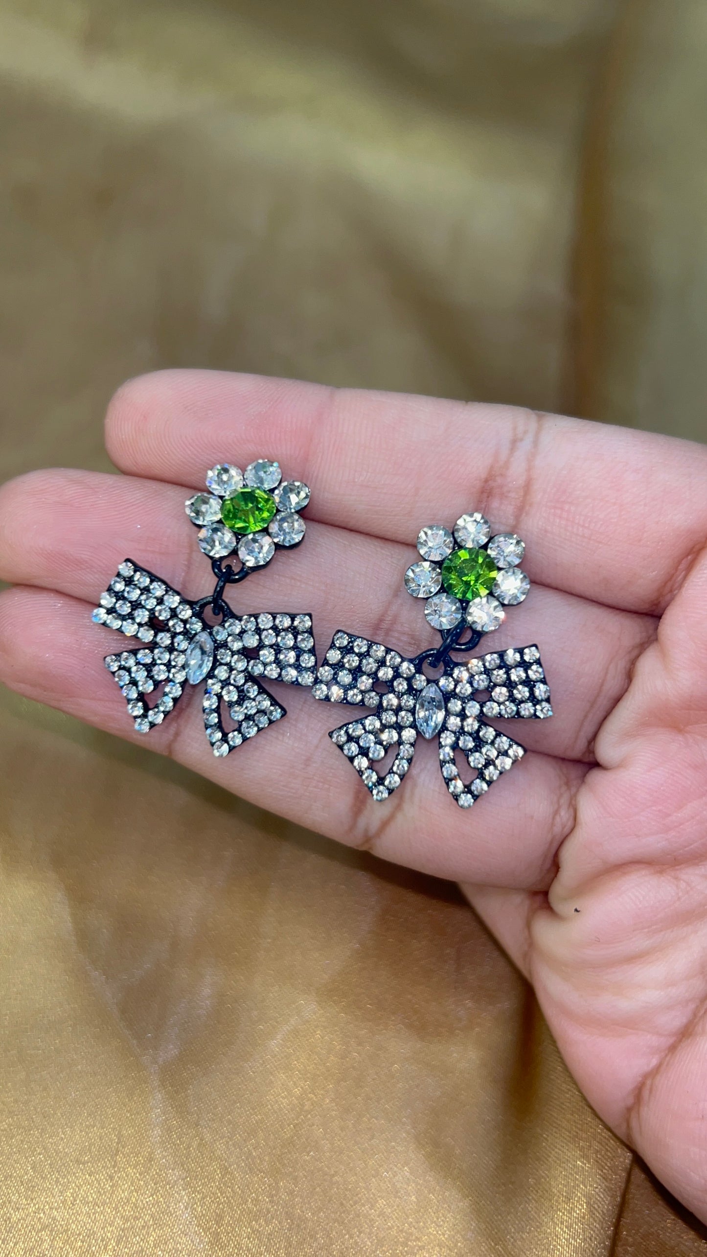 Pair of decorative earrings with green gemstones held in a hand against a beige background