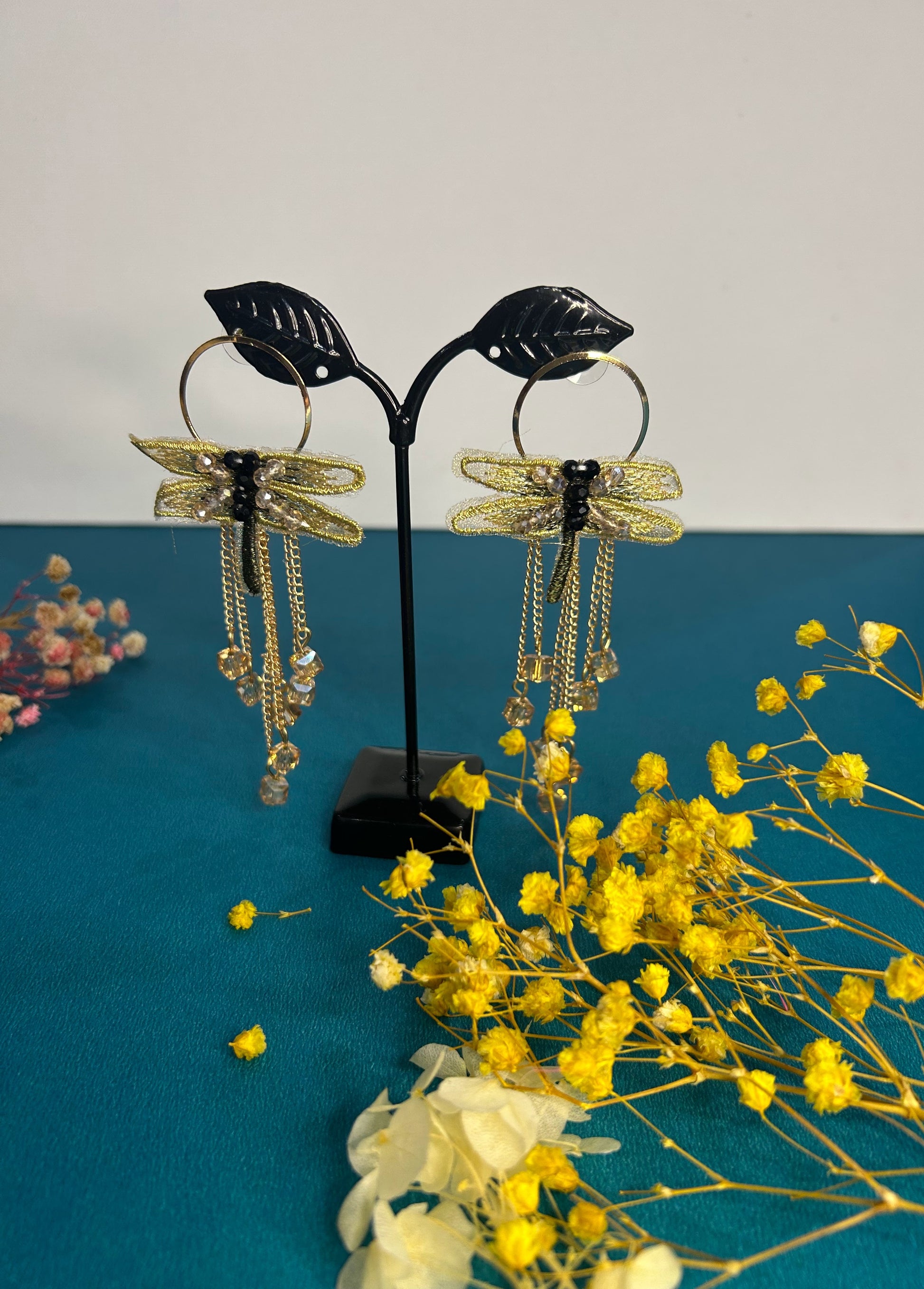 Gold dragonfly earrings on a stand with yellow flowers on a blue surface