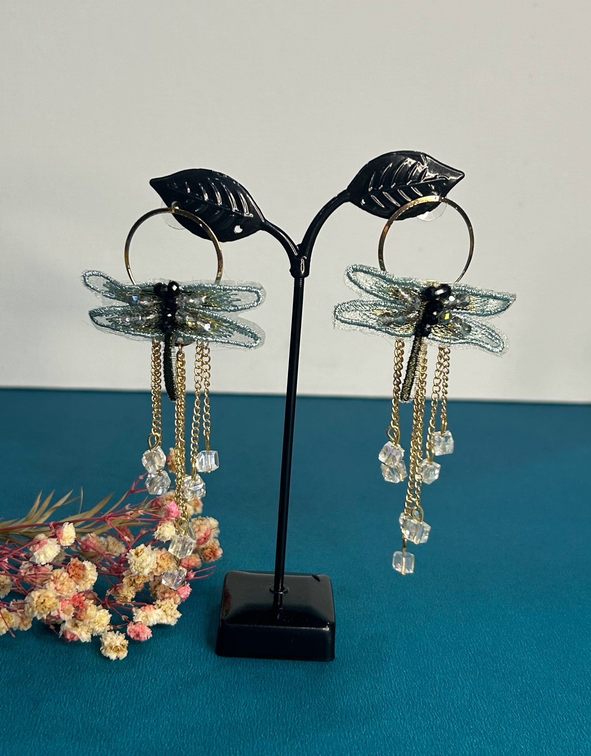 Dragonfly-shaped earrings on a black stand with a floral arrangement on a blue surface.