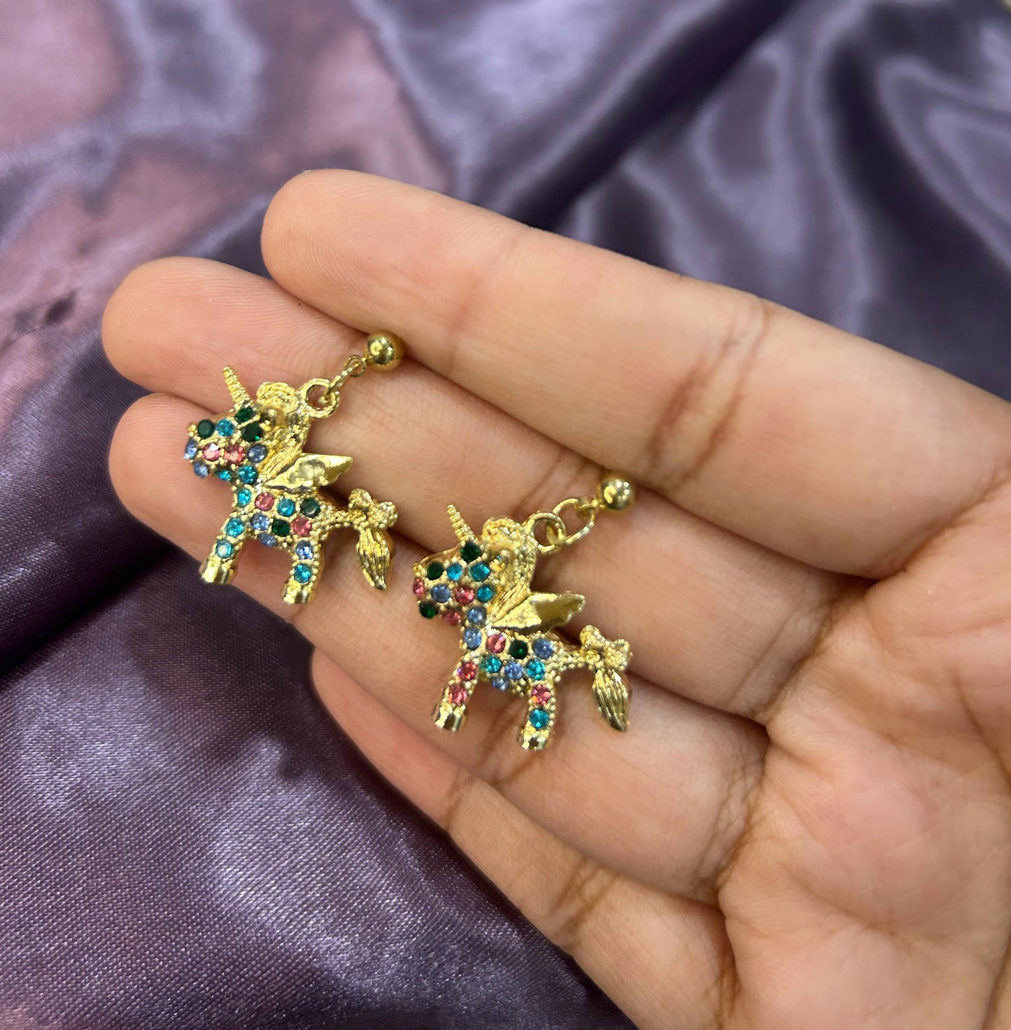 Gold unicorn charms with colorful gemstones held in a hand against a purple fabric background