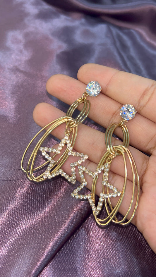 Gold hoop earrings with star designs held by a hand on a purple satin background