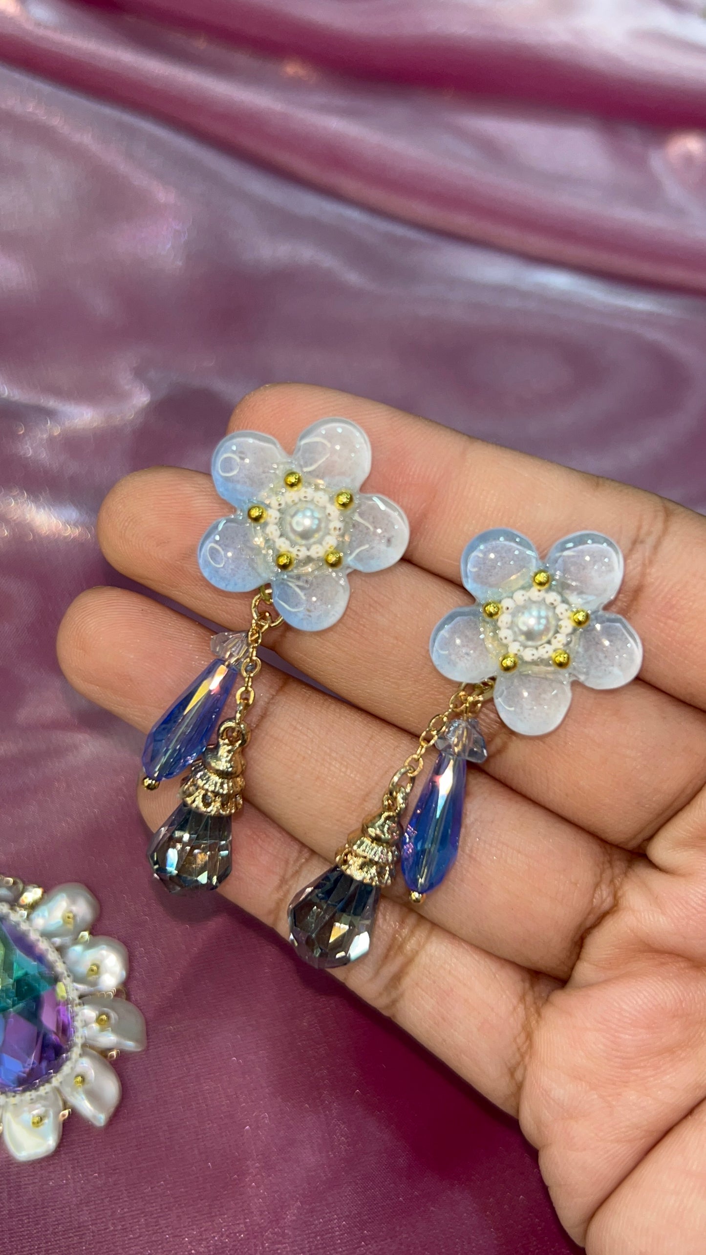 Flower-shaped earrings with gemstones on a hand against a pink background