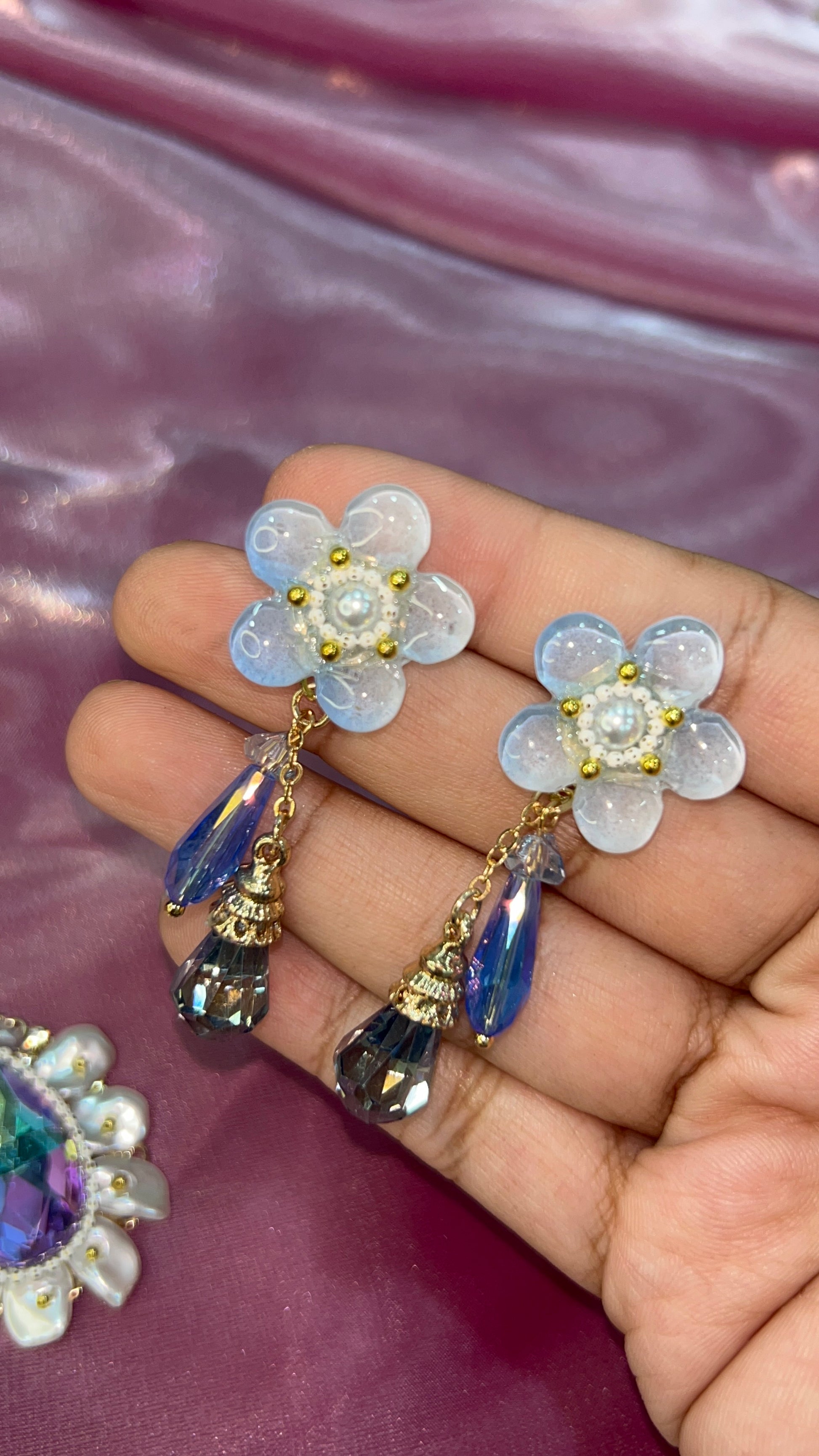 Flower-shaped earrings with gemstones on a hand against a pink background
