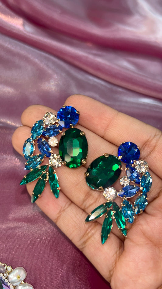 Decorative brooch with green and blue gemstones held in a hand against a pink fabric background