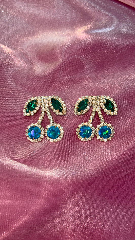 Cherry-shaped earrings with green and blue gemstones on a pink satin background