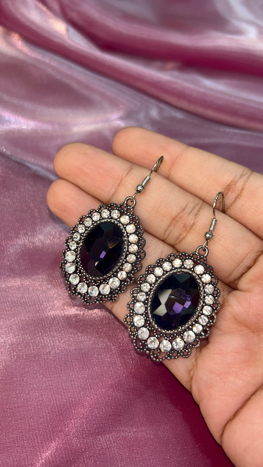 Pair of earrings with purple gemstones held on a hand against a pink fabric background