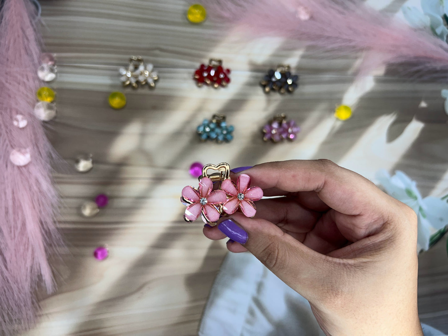 Hand holding a pink floral hair clip with colorful earrings in the background