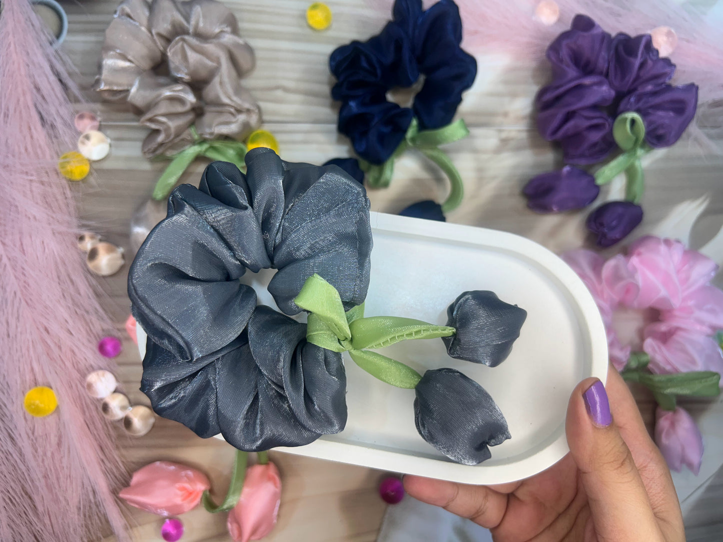 Gray scrunchie with green ribbon held over a tray with more scrunchies and flowers.