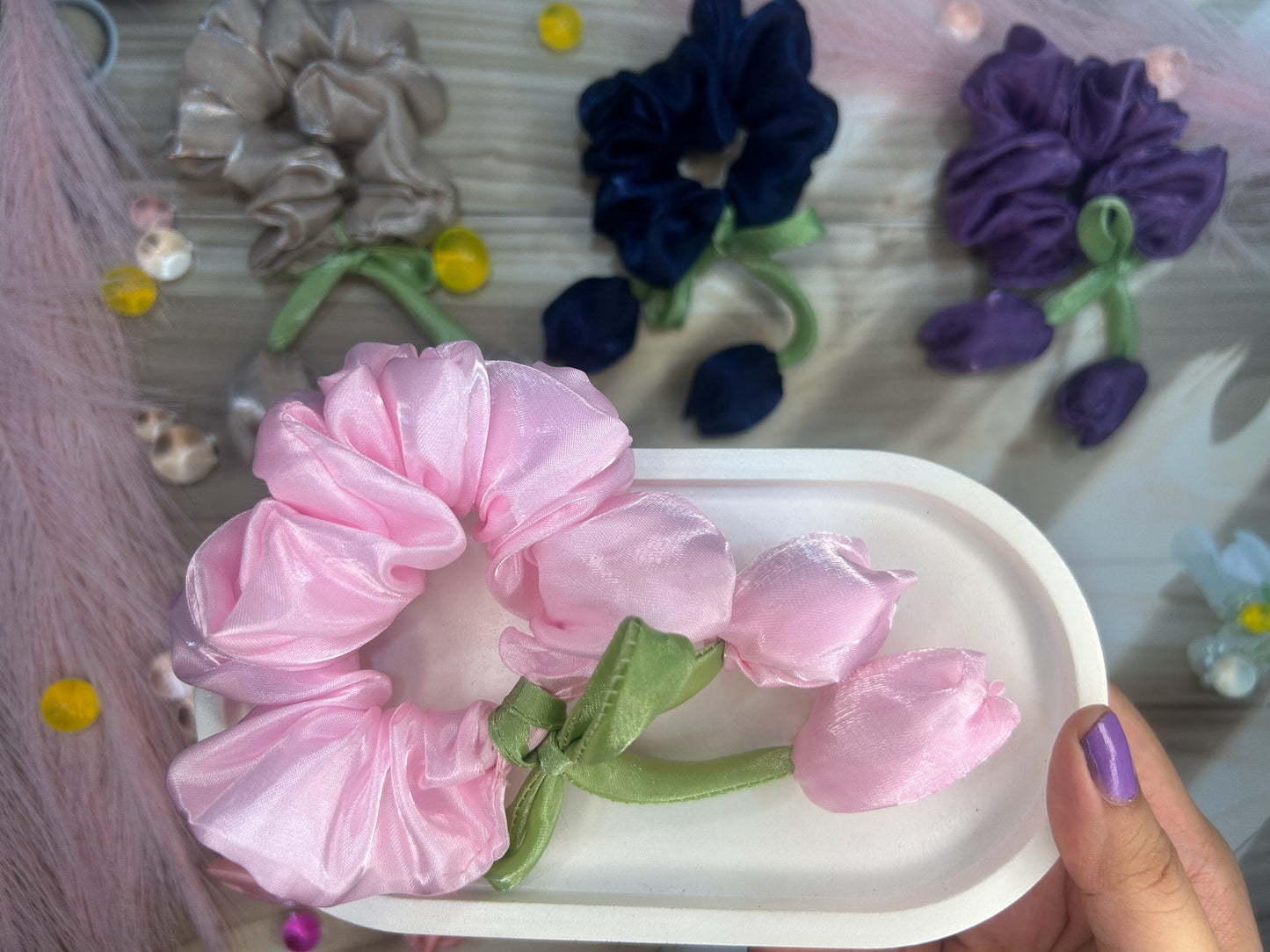 Pink scrunchie with green leaves on a white tray, surrounded by colorful flowers.