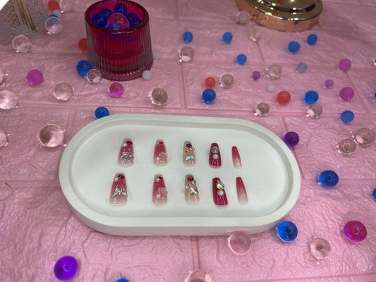 Set of artificial nails in a white tray on a pink surface with decorative gems.