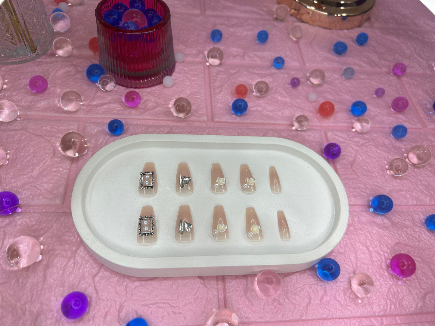Set of false nails in a white tray on a pink surface with decorative gems.