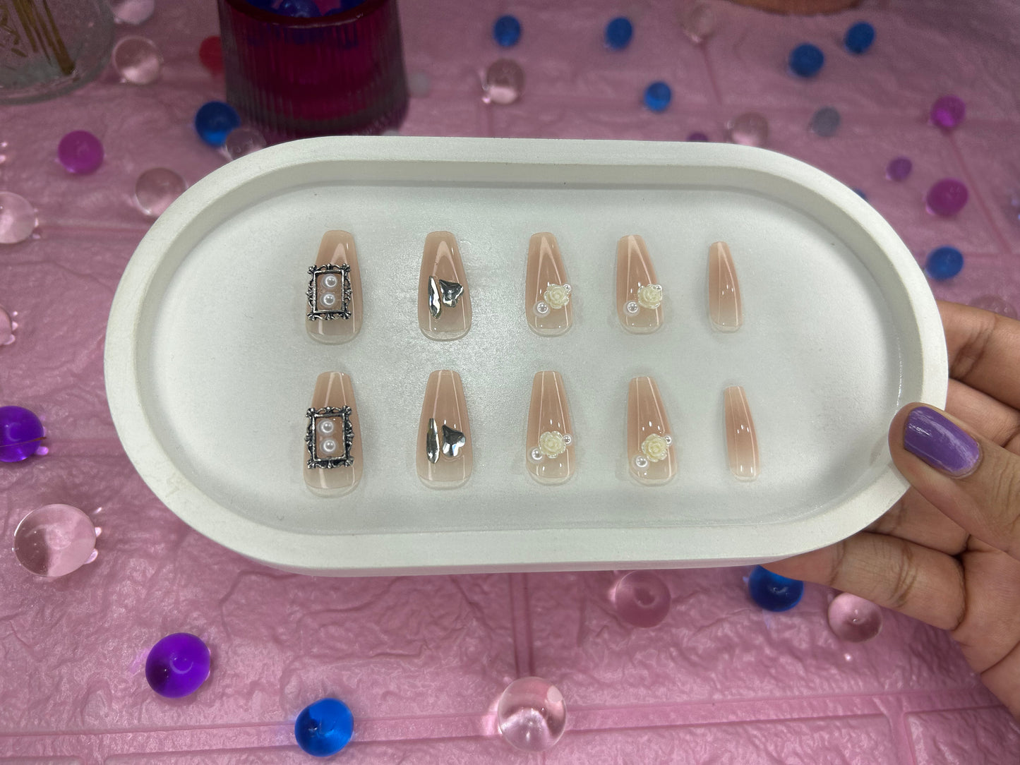 Tray of nail designs held by a hand on a pink surface with decorative items.