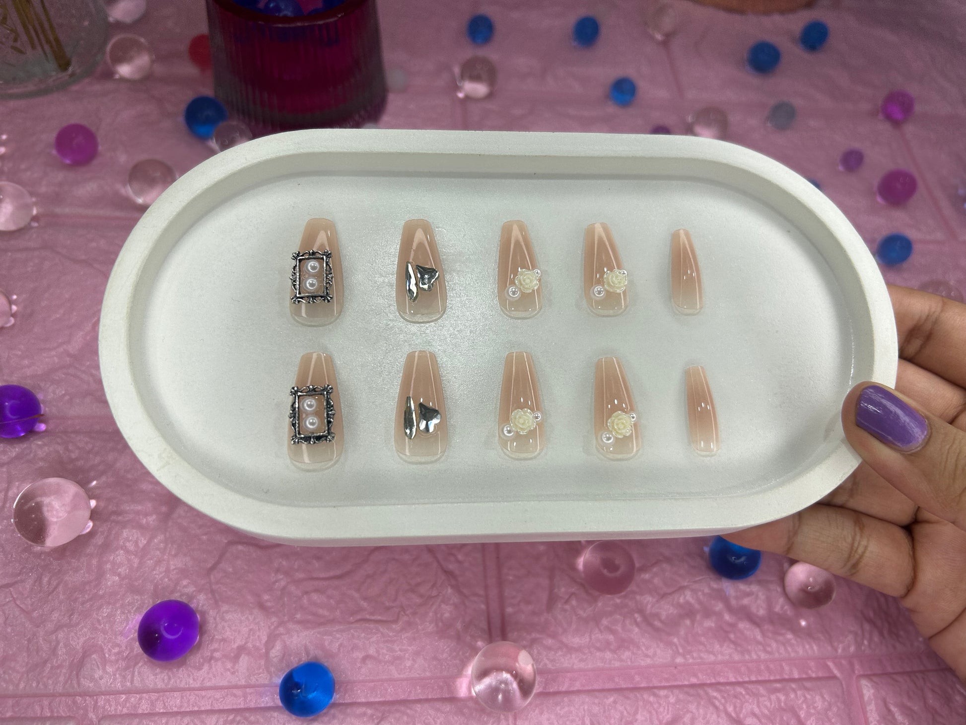 Tray of nail designs held by a hand on a pink surface with decorative items.