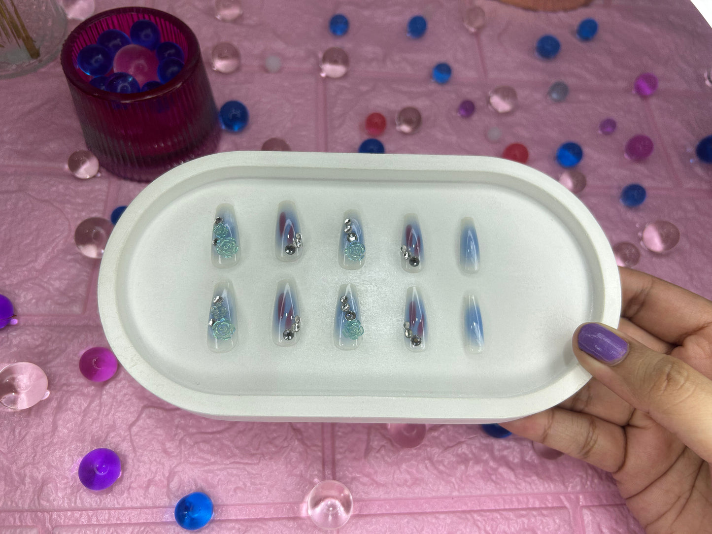 Set of decorative nail tips in a white case held by a hand on a pink surface with colorful beads.