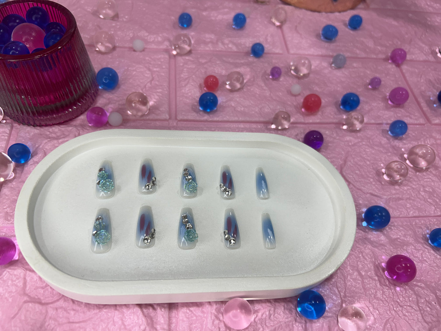 Set of nail art samples in a white tray on a pink surface with decorative beads.