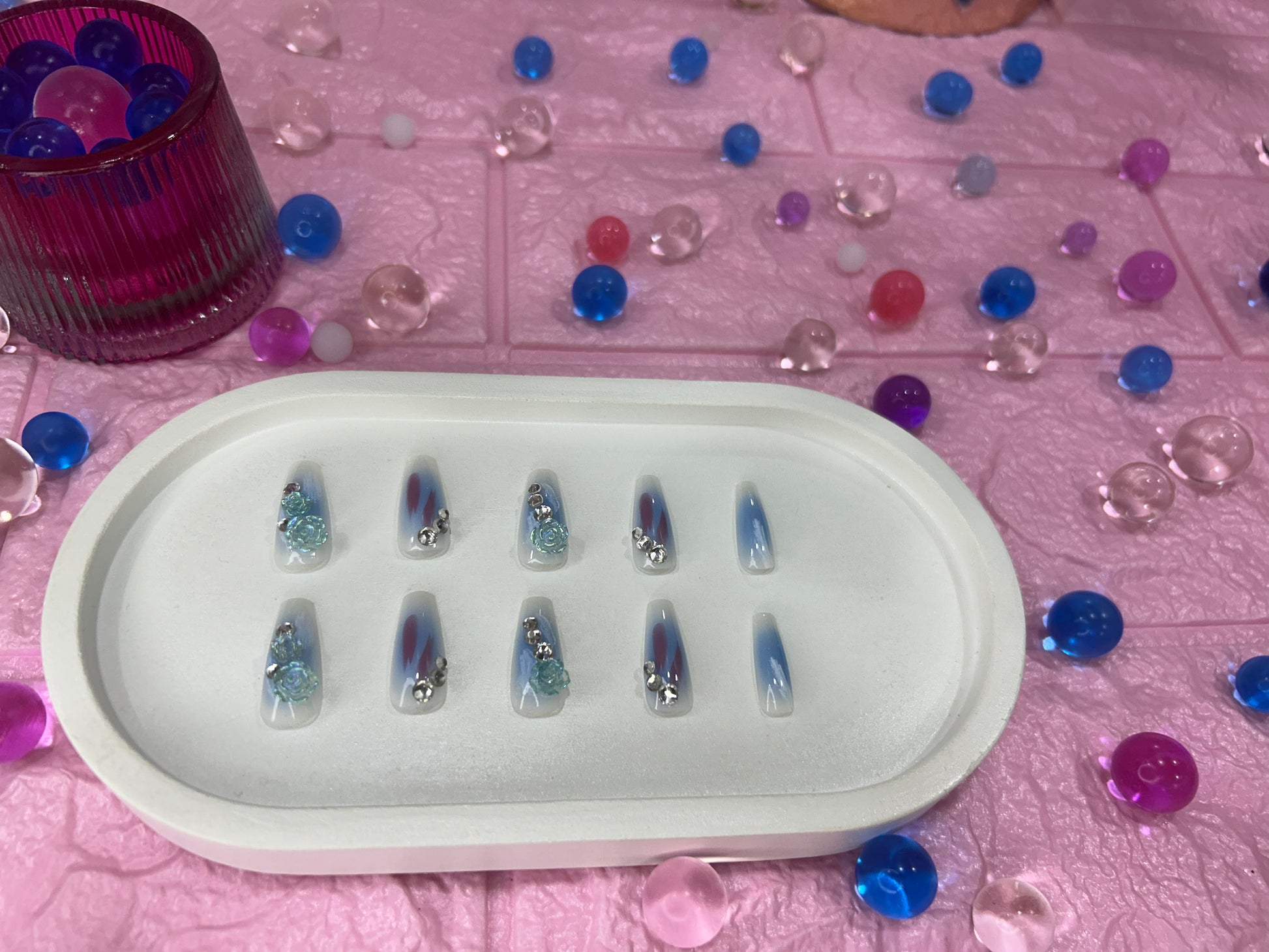 Set of nail art samples in a white tray on a pink surface with decorative beads.