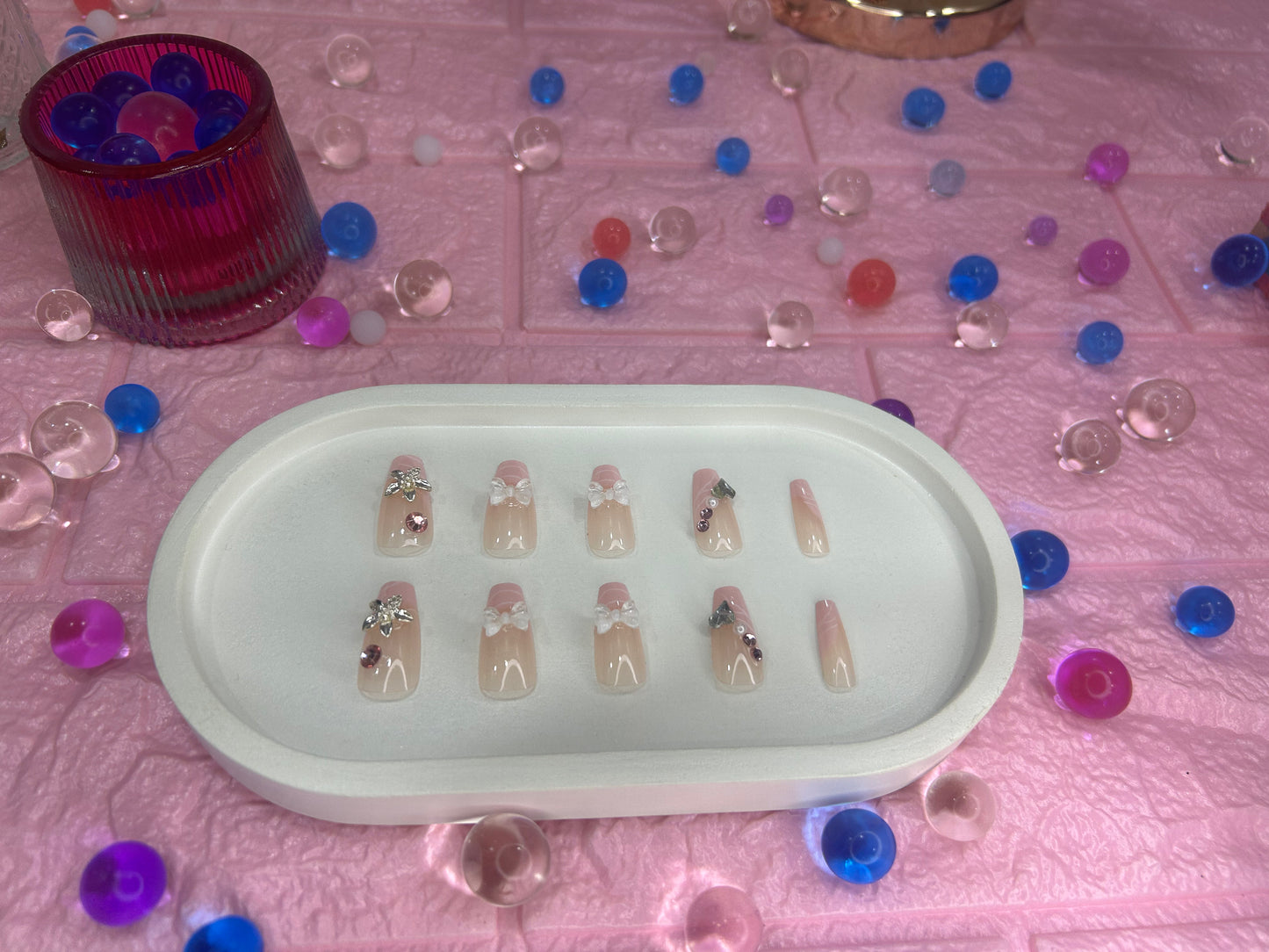 Set of false nails in a white tray surrounded by colorful buttons on a pink textured surface