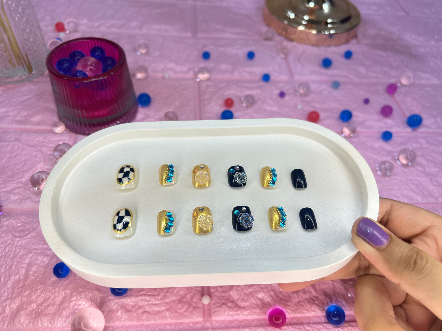 Set of nail extensions in a white tray held by a hand on a pink surface with decorative items.