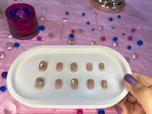 Set of false nails in a white tray with a hand holding them, on a pink surface with decorative elements.