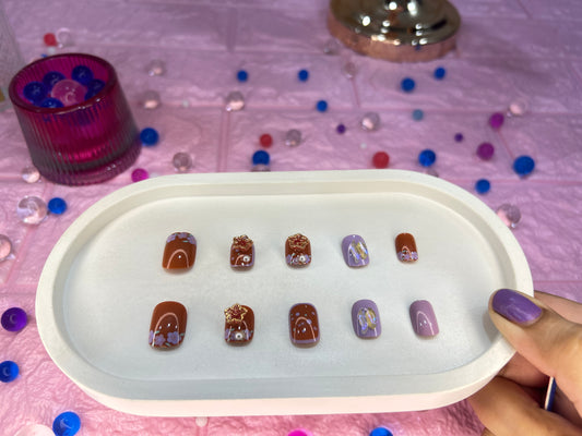 Set of miniature brown and purple fake nails on a white tray with colorful beads in the background.