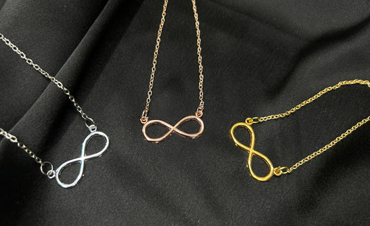Three infinity symbol necklaces in silver, rose gold, and yellow gold on a black fabric background.