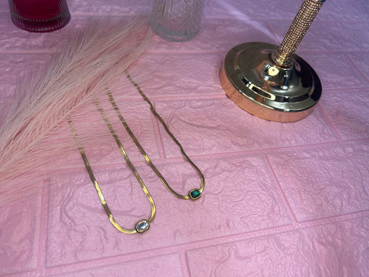 Two gold necklaces with green gemstones on a textured pink surface.