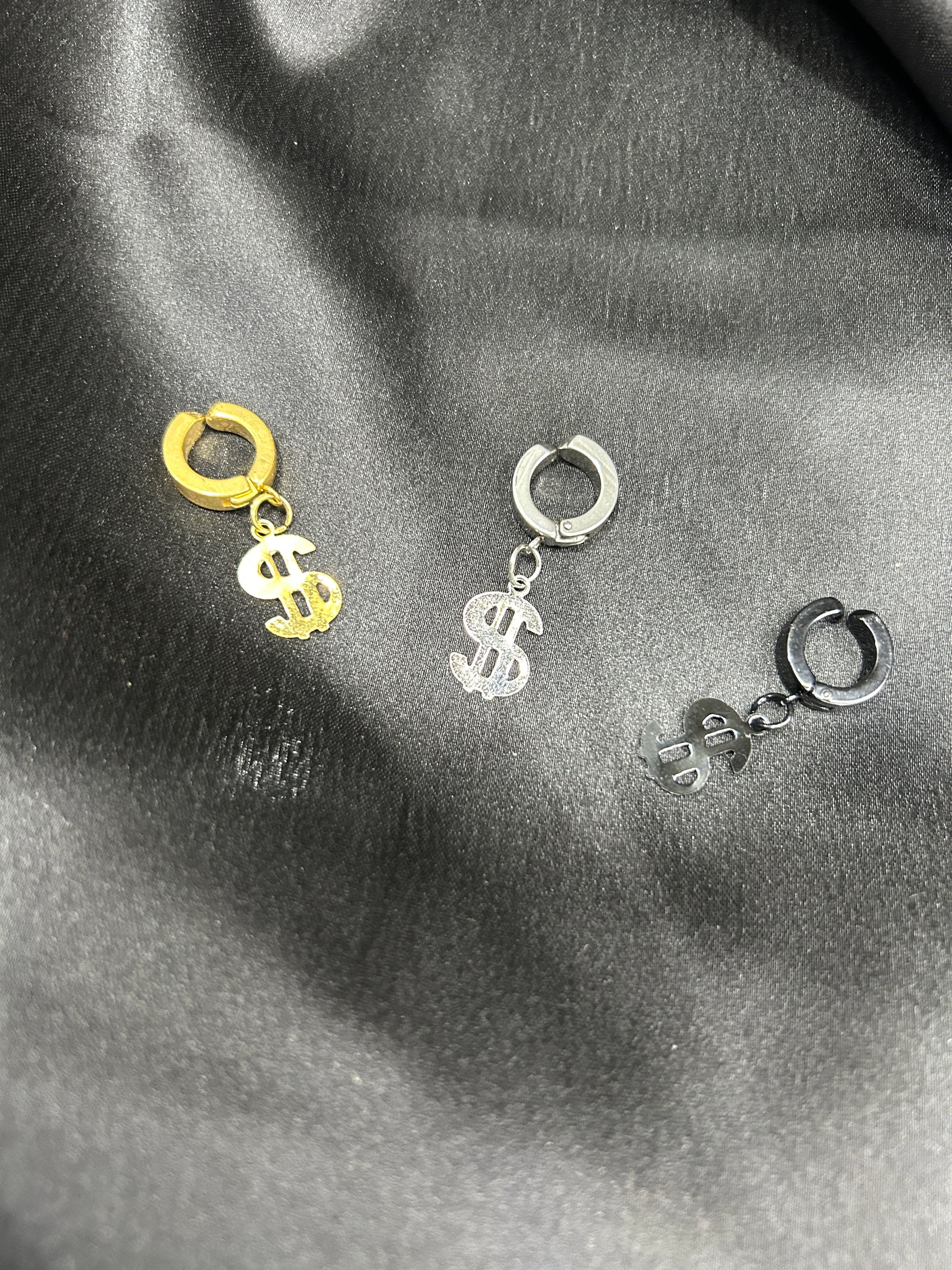 Two pairs of earrings with dollar sign charms on a dark fabric background