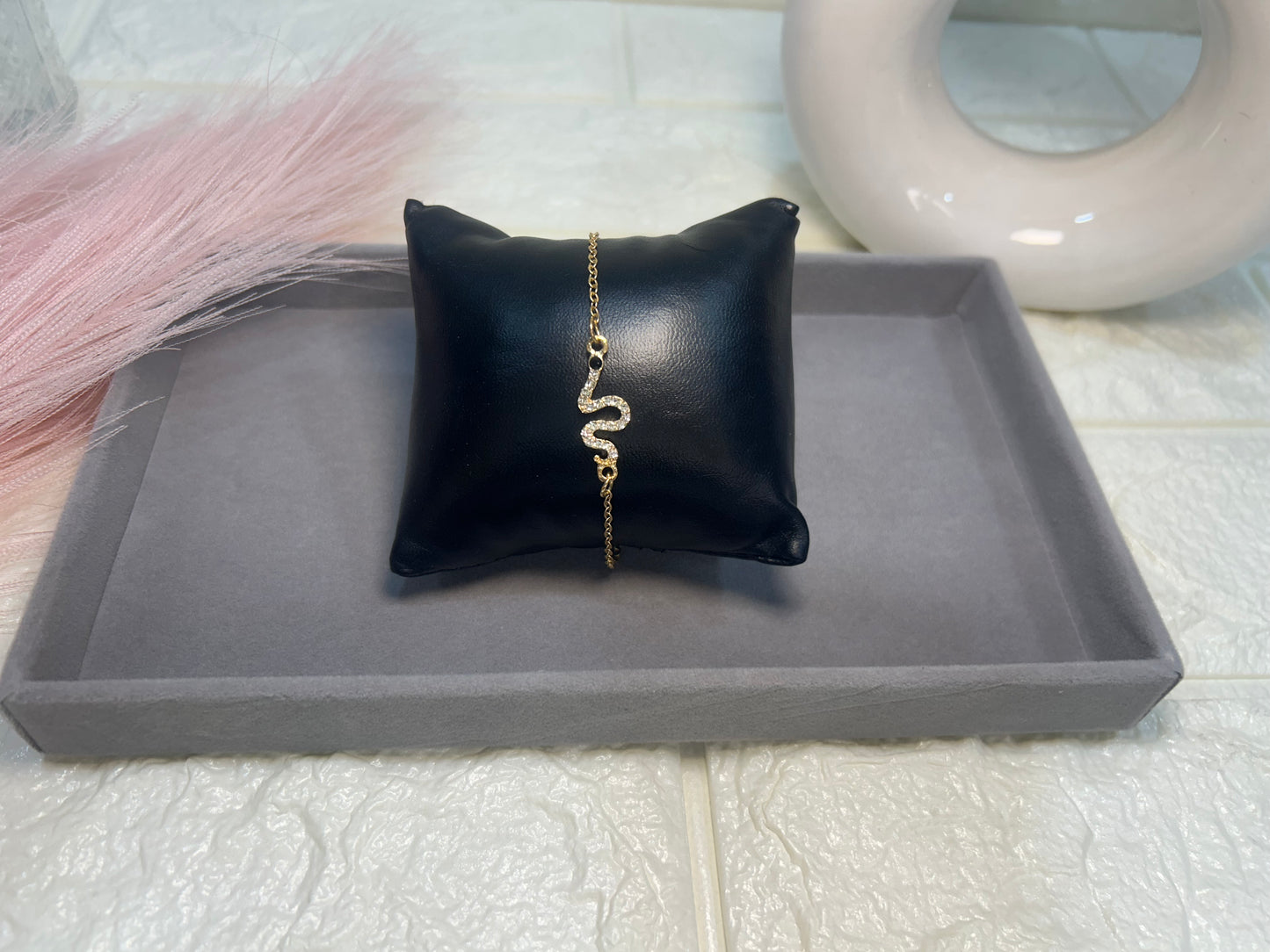 Gold bracelet on a black pillow with a gray tray underneath, on a light pink surface.