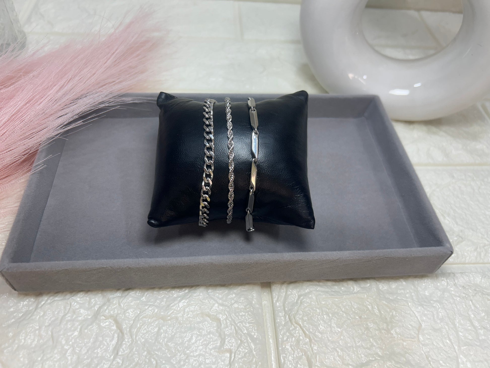 Two silver bracelets on a black pillow with a gray tray on a textured white surface.