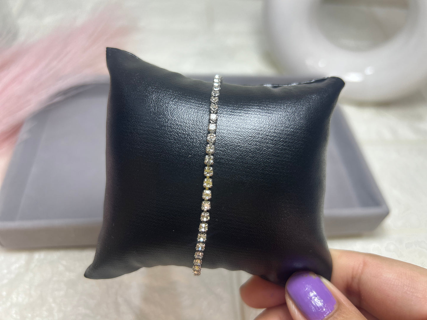 Bracelet with clear stones on a black leather pillow held by a hand with purple nail polish.